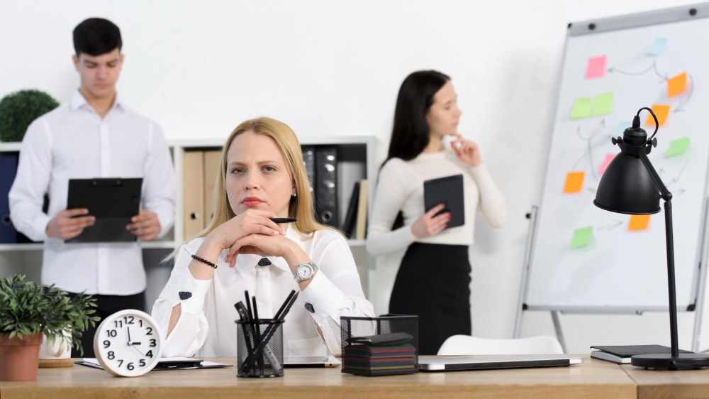 Businesswoman depicting unconscious bias in the workplace seated at a desk with a thoughtful expression, while colleagues work in the background